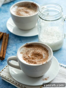 A close-up shot of a Turmeric Latte in a white mug, garnished with cinnamon, beside a few tea bags and fresh turmeric root.