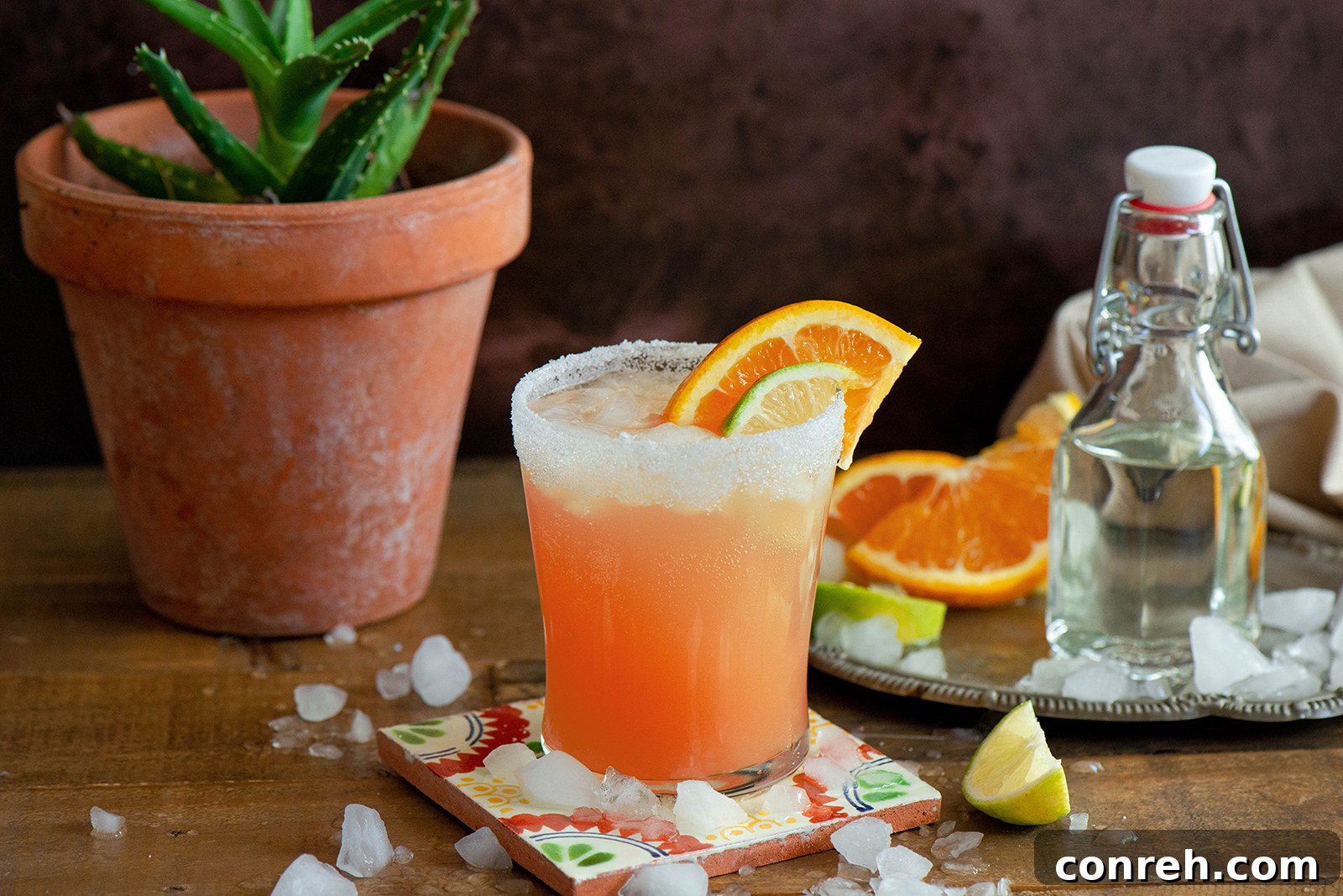 A Paloma cocktail in a sugar-rimmed glass, with a lime wedge garnish, next to fresh grapefruit and a shaker.
