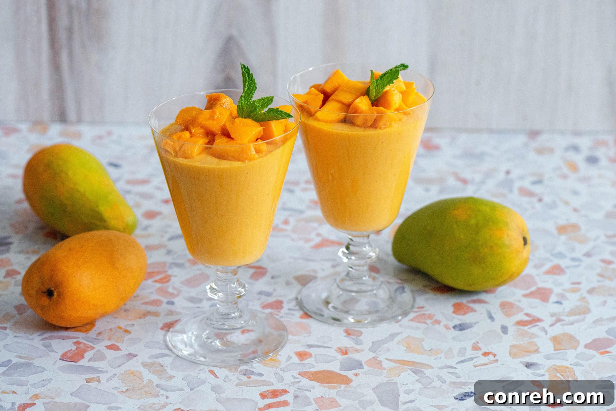 Two glasses of mango mousse garnished with mint leaves, next to a bowl of fresh mango chunks.