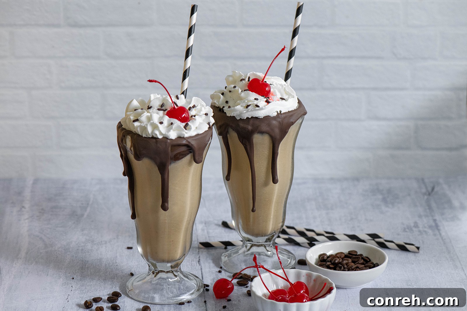 Two glasses of Mexican Coffee Milkshakes, garnished with whipped cream, chocolate syrup, and cherries, on a wooden surface.