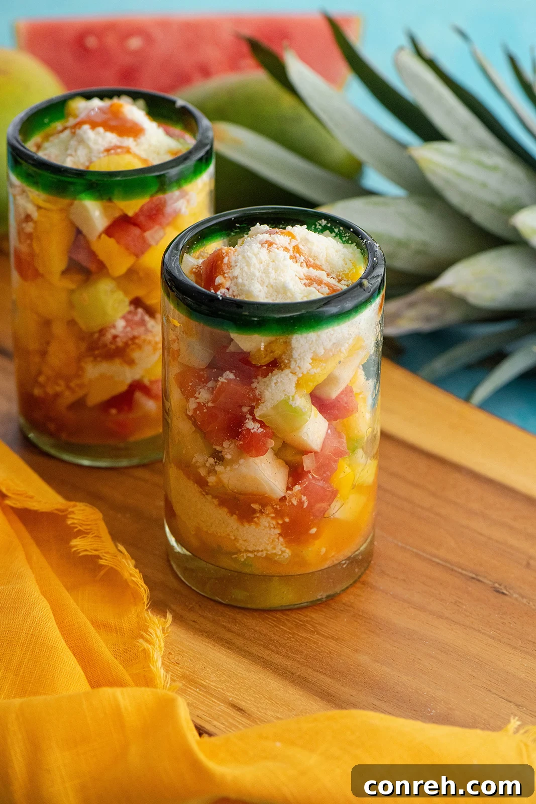 Morelia's Refreshing Gazpacho 2 A tall glass filled with vibrant Gazpacho Moreliano, showcasing layers of fresh fruit, onion, cotija cheese, and a drizzle of El Yucateco hot sauce, ready to be enjoyed as a light treat.