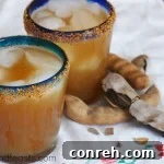 A refreshing glass of Agua de Tamarindo garnished with a chili salt rim, beside fresh tamarind pods.